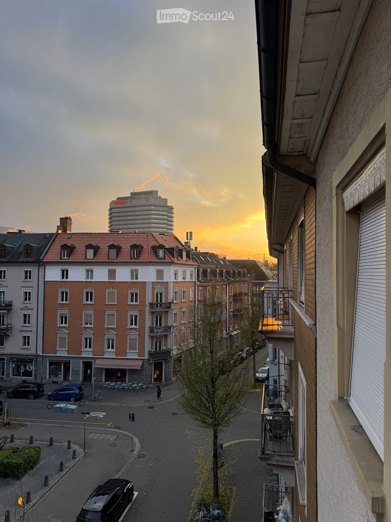 Urban street scene at sunset with multi-story buildings, parked cars, and a visible balcony with outdoor furniture.