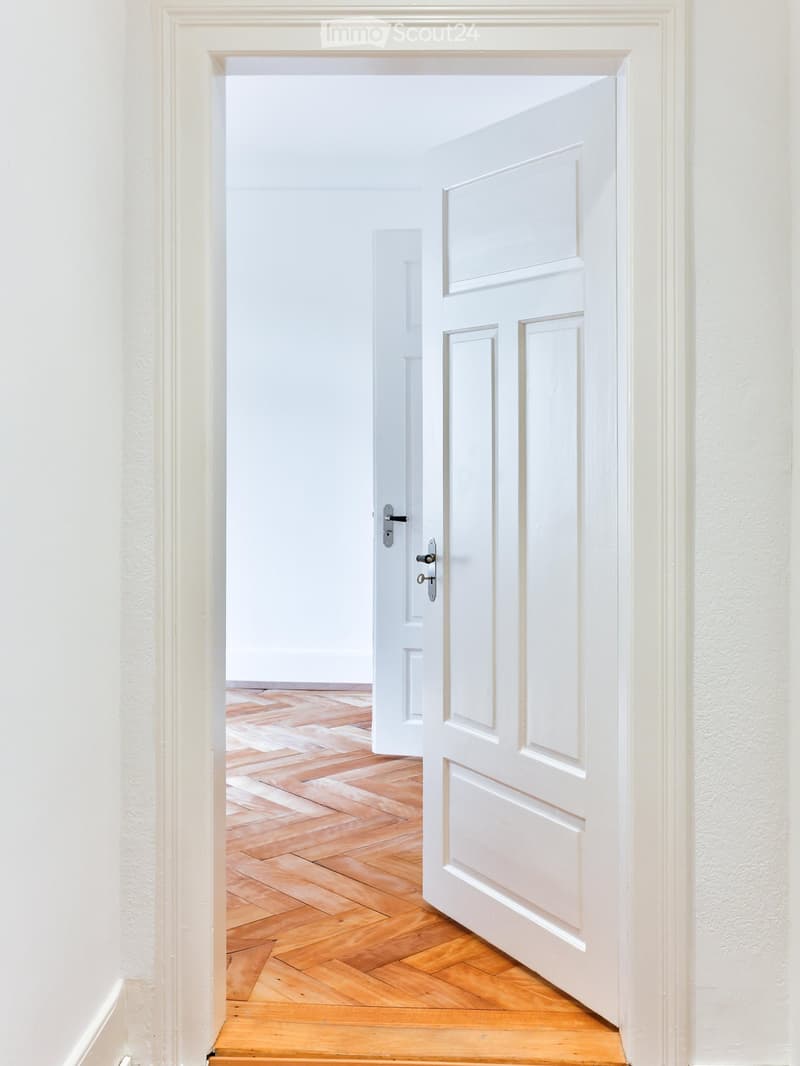 A clean, white-walled hallway with a wooden herringbone floor, leading to an open door that reveals another room.