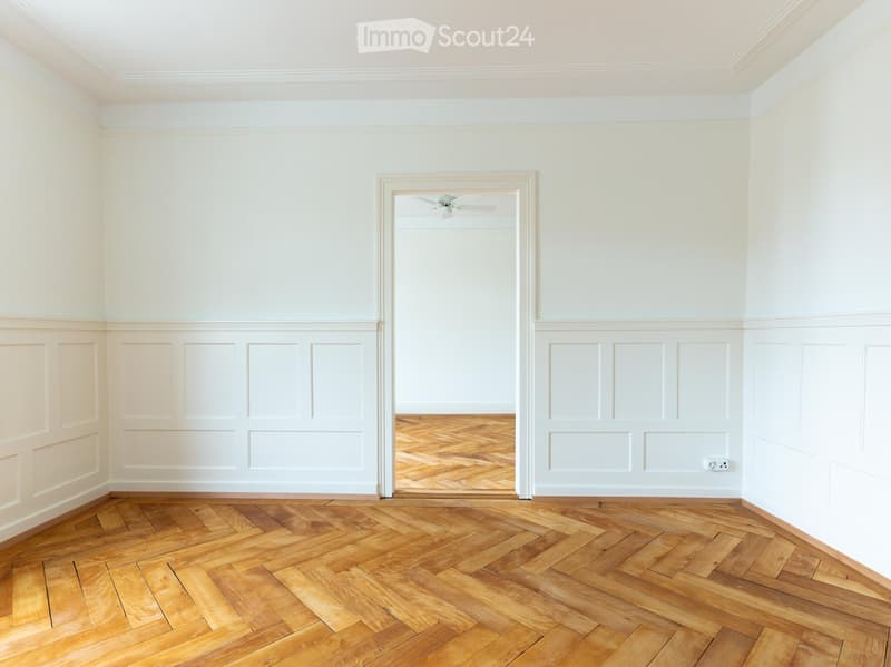 Empty room with light-colored walls, wooden herringbone flooring, and a white ceiling with a visible light fixture. The room features decorative paneling on the lower half of the walls and a doorway leading to another room.