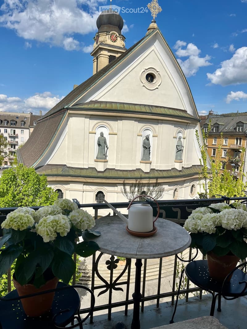 Balcony with a round table, two chairs, and potted flowers, overlooking a historic church with a dome and clock tower.