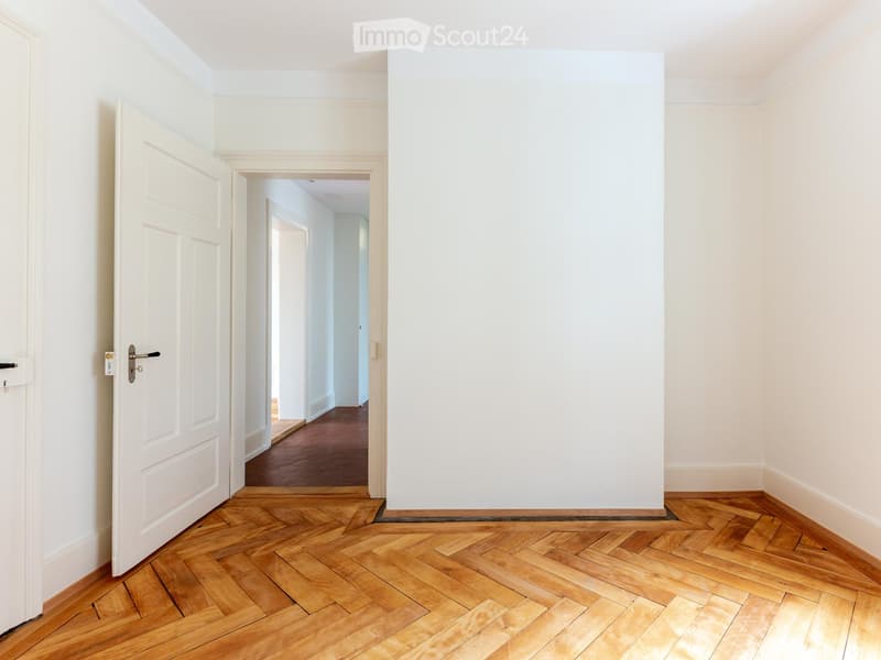Empty hallway with white walls, wooden herringbone flooring, and an open door leading to another room.
