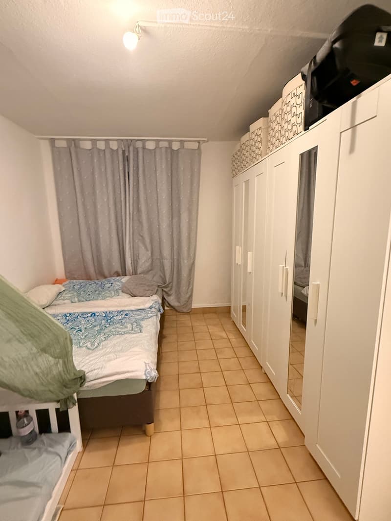 Cozy bedroom with a single bed, a wardrobe, and a curtain covering a window. The room features tiled flooring and white walls.