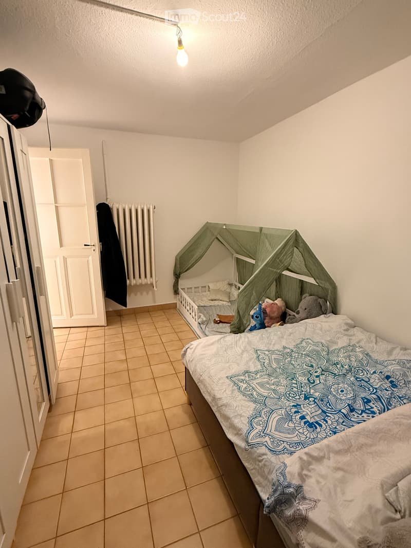 A small bedroom with a single bed, a toy tent, and several stuffed toys on the bed. The room has tiled flooring and white walls, with a radiator and a partially open door visible.