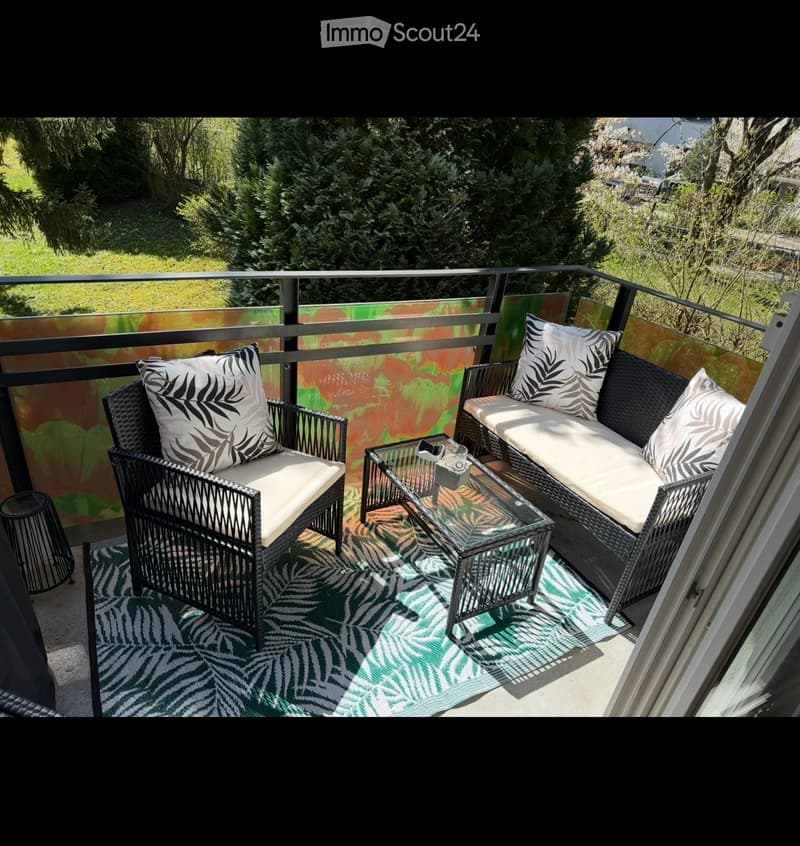 Balcony with a sofa, chair, glass coffee table, and a small stool, surrounded by greenery.