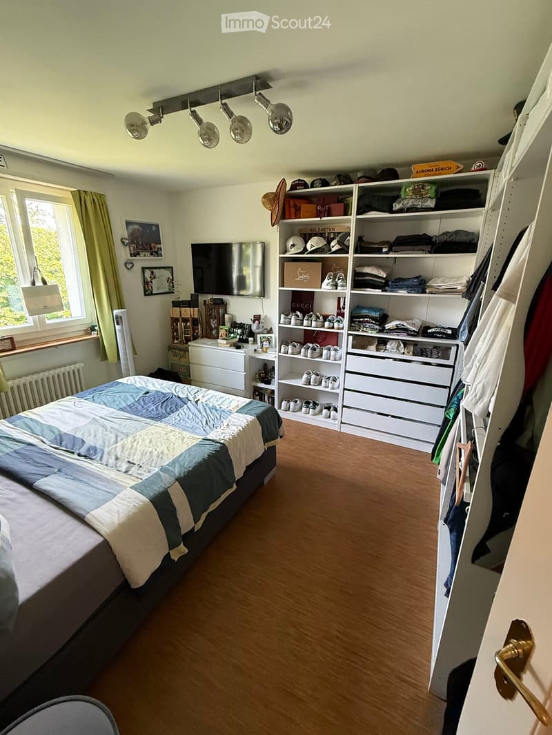 Well-lit bedroom with wooden flooring, a large bed with a blue and white quilt, a mounted TV, built-in wardrobe with shelves and drawers, and a window with green curtains.