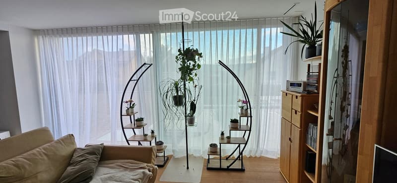 A modern living room with a beige sofa, a tall plant stand with multiple shelves holding small potted plants, a curved floor lamp, a wooden bookshelf with books and a radio, and a large window with sheer curtains.