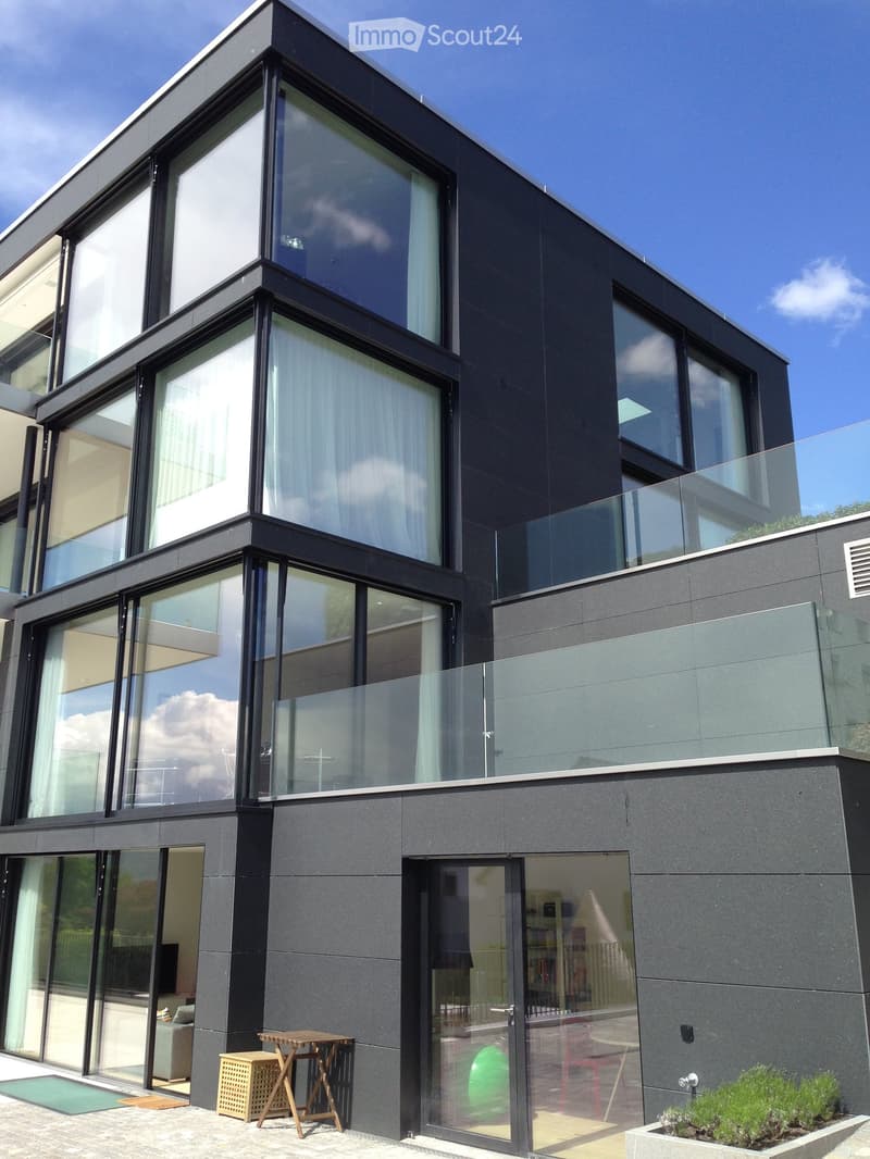 Modern two-story house with dark grey exterior, large glass windows, and a balcony with glass railing. The ground floor features a small outdoor seating area with a table and chairs.