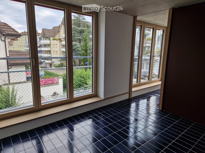 Empty room with large windows and black tiled floor, overlooking a residential area with buildings and greenery.