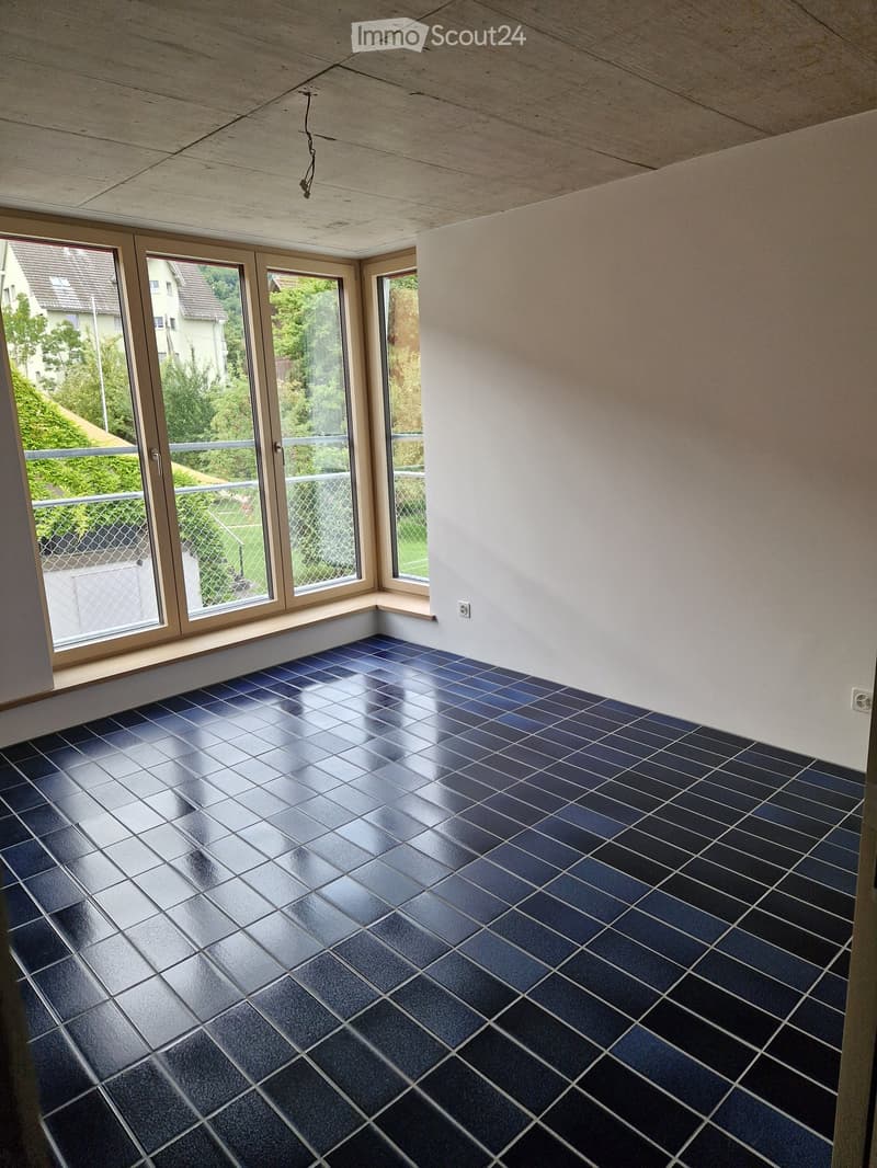 Empty room with dark blue tiled floor and large windows offering a view of greenery outside. The walls are white and the ceiling is light-colored.