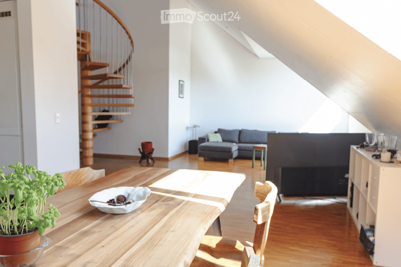 Bright, open living and dining area with wooden flooring, a grey sectional sofa, a wooden dining table with chairs, a small green stool, a potted plant, and a decorative bowl on the table. A modern staircase leads upwards.