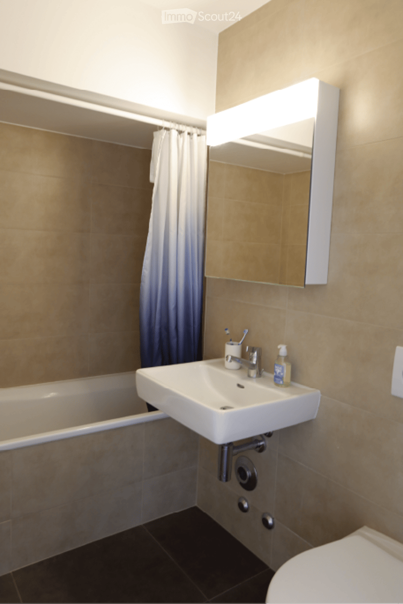 Modern bathroom with a white sink, illuminated mirror, toothbrush holder, and a bathtub with a shower curtain.