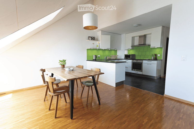 Bright, modern kitchen and dining area with white cabinets, green backsplash, wooden dining table, and chairs. Features a hanging pendant lamp and a potted plant on the table.