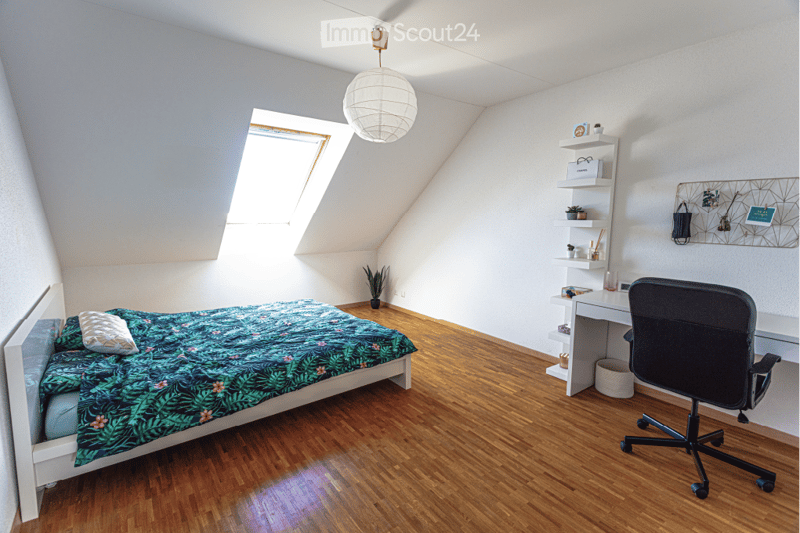 Bright bedroom with wooden flooring, a single bed with a green and pink patterned duvet, a desk with a chair, and a shelf with decorative items.