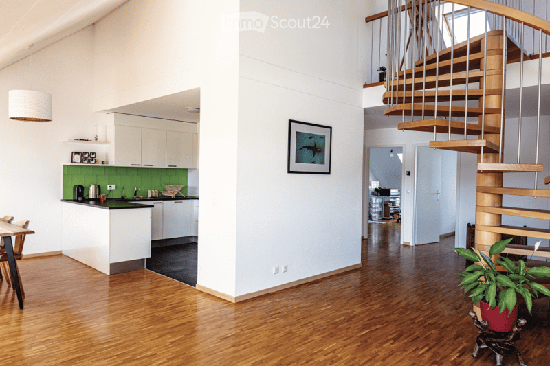 Open-plan living and kitchen area with light-colored cabinets, green backsplash, wooden flooring, a staircase with wooden steps and metal railings, and a dining table with chairs.