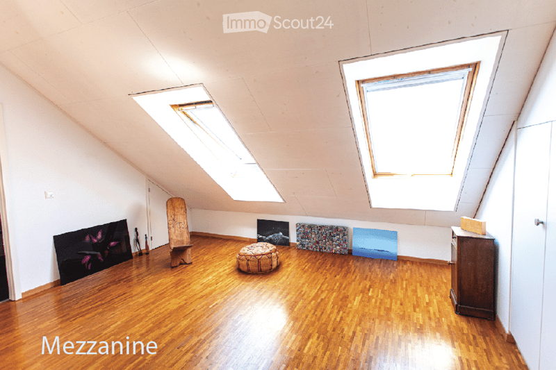 Spacious attic room with wooden flooring, skylights, and minimal furnishings including a round stool, a small cabinet, and several artworks on the walls.