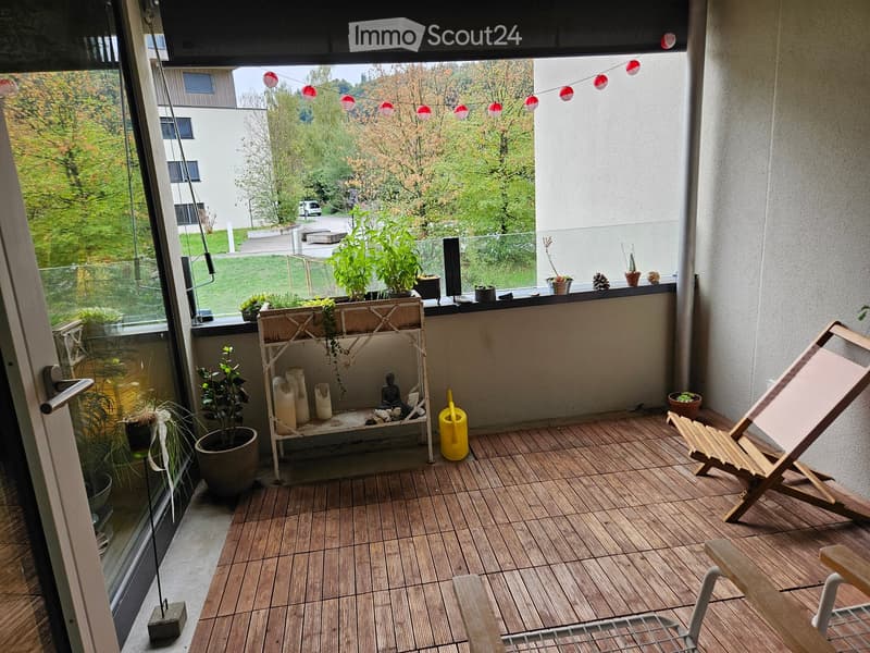 Balcony with wooden decking, potted plants, a small shelf with candles and a statue, a yellow watering can, and a wooden folding chair.