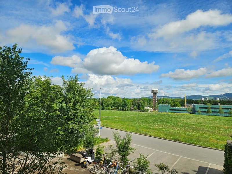 A grassy field with trees and a modern building in the background, under a partly cloudy sky. There are parked bicycles and a scooter in the foreground.