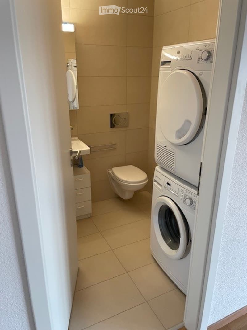 Modern bathroom with beige tiles, a toilet, sink, mirror, cabinet, and stacked washing machine and dryer.