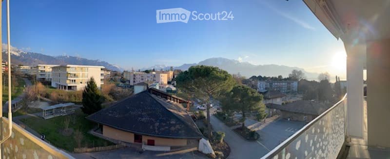Balcony overlooking a scenic town with mountains in the background, featuring a railing and a view of buildings and greenery.