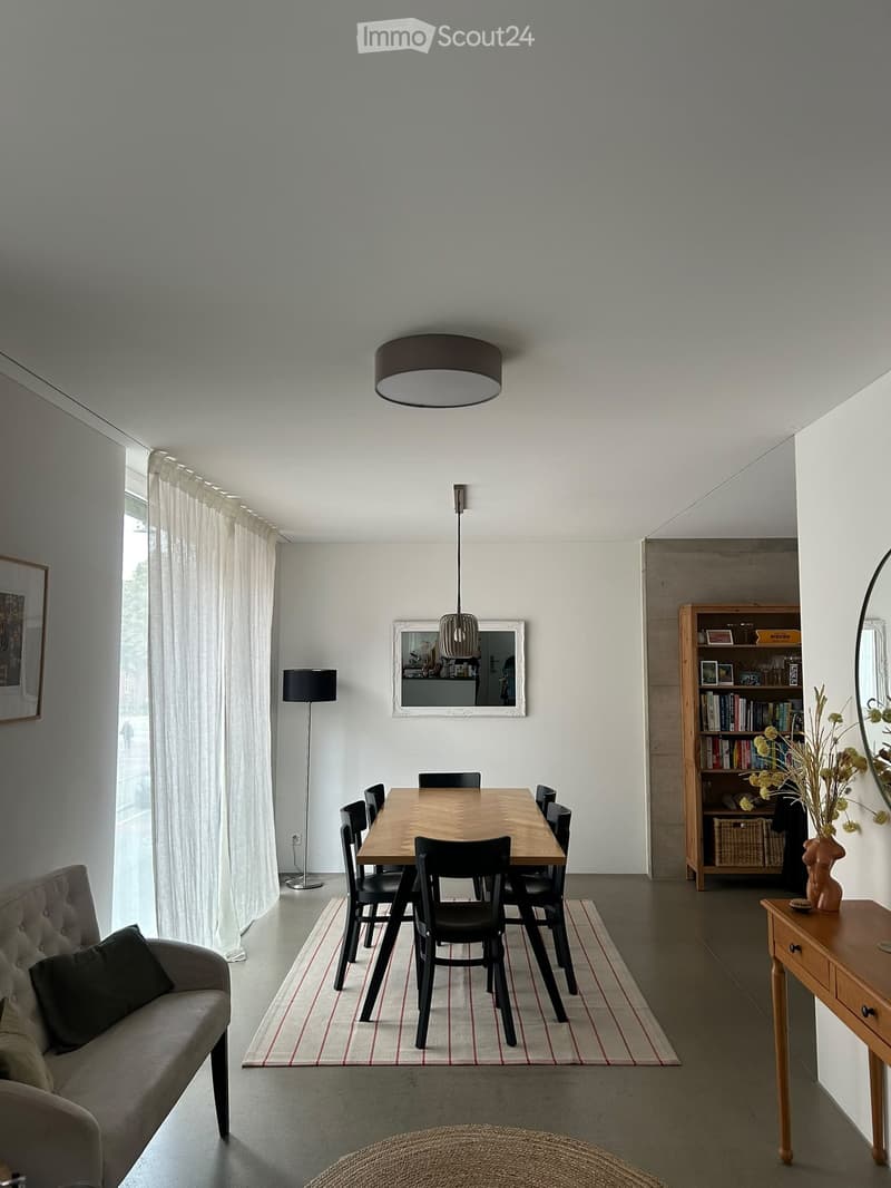 Modern dining and living area with a wooden dining table, black chairs, a grey sofa, bookshelf, floor lamp, and decorative items.