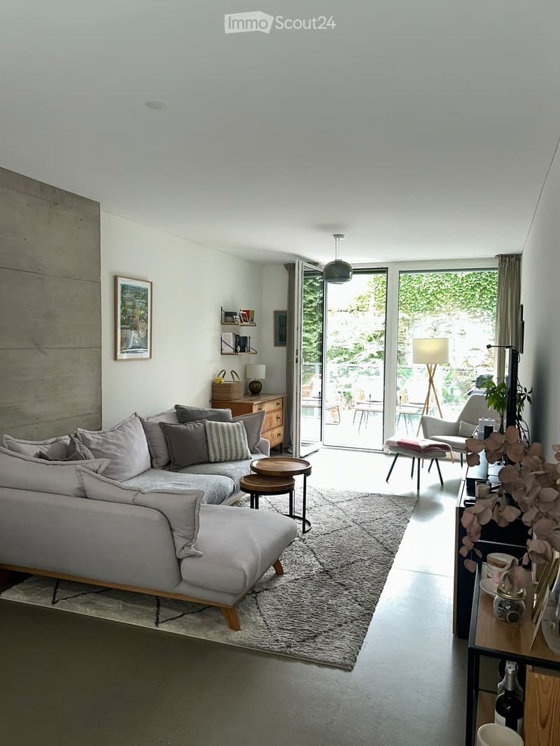 Modern living room with grey sectional sofa, wooden coffee table, side tables, and a view to a balcony or terrace area with outdoor furniture.