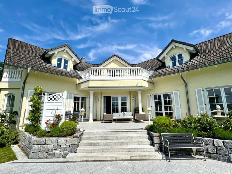 Large house, 3 stories, brown roof, white facade, multiple windows, balcony with railing, patio with seating, stone steps, garden with plants and fountain, driveway