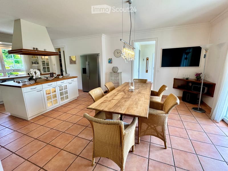 Dining room with wooden table, wicker chairs, white kitchen with island, refrigerator, chandelier, TV, and decorative items