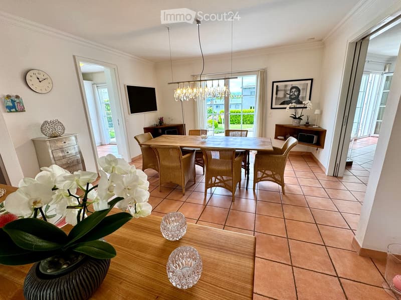 Dining room with wooden table, chairs, chandelier, TV, picture frame, clock, flower vase, cabinet, and tiled floor.
