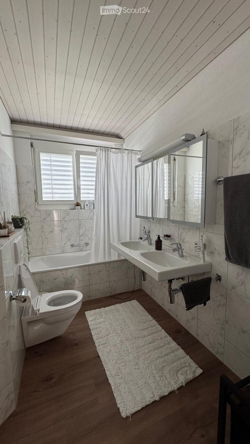 Modern bathroom with marble tiles, double sink, toilet, bathtub, large mirror, and a white shag rug on the floor.