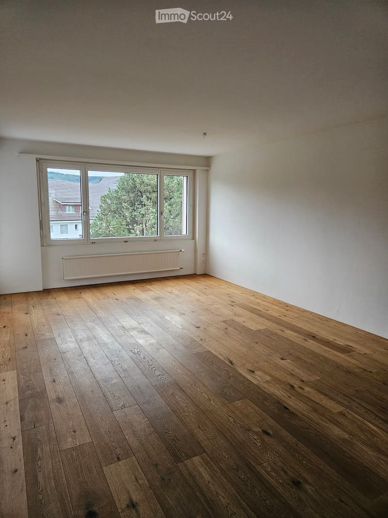 Empty room with wooden flooring, white walls, and a large window with a view of trees and a neighboring house.