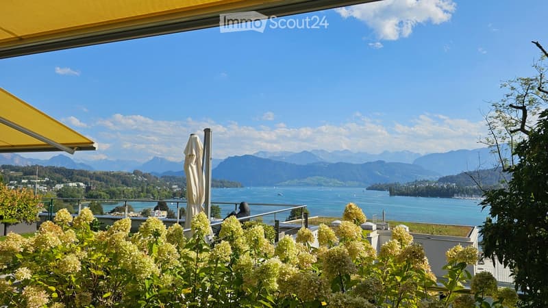 Balcony with a view of a lake and mountains, featuring a yellow awning, a closed umbrella, and flowering shrubs in the foreground.