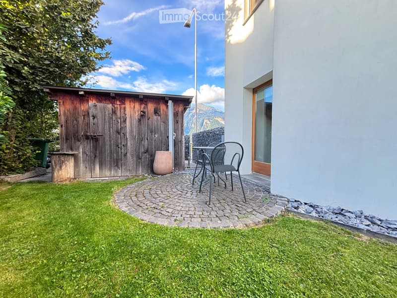 A small outdoor patio area with a table and chairs, surrounded by a stone pathway, grassy lawn, and a wooden shed.