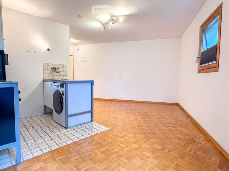 A small, well-lit room with wooden flooring, a washing machine, and a tiled area for the machine. The room has white walls, a window with blinds, and recessed lighting.