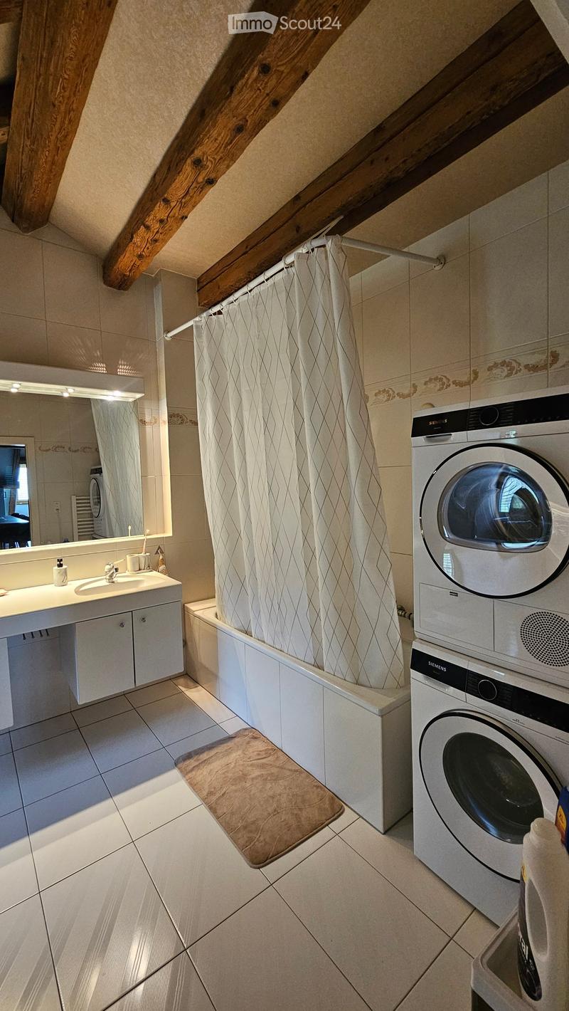 Bathroom with a sink, mirror, bathtub, washing machine, and shower curtain. The room features exposed wooden beams on the ceiling and tiled walls.