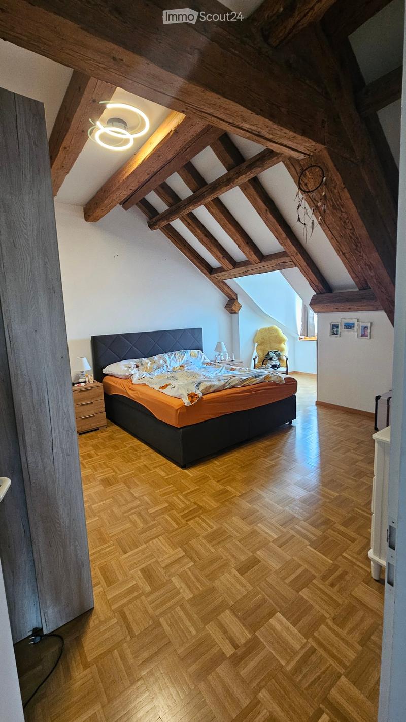 Cozy attic bedroom with wooden beams, herringbone wooden flooring, a large bed with orange bedding, a nightstand with a lamp, and a small seating area with a chair and dreamcatcher.