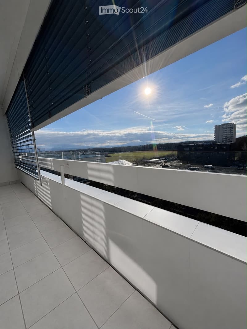 Modern balcony with white tiled flooring, white railings, and large glass windows offering a view of a cityscape and distant mountains.