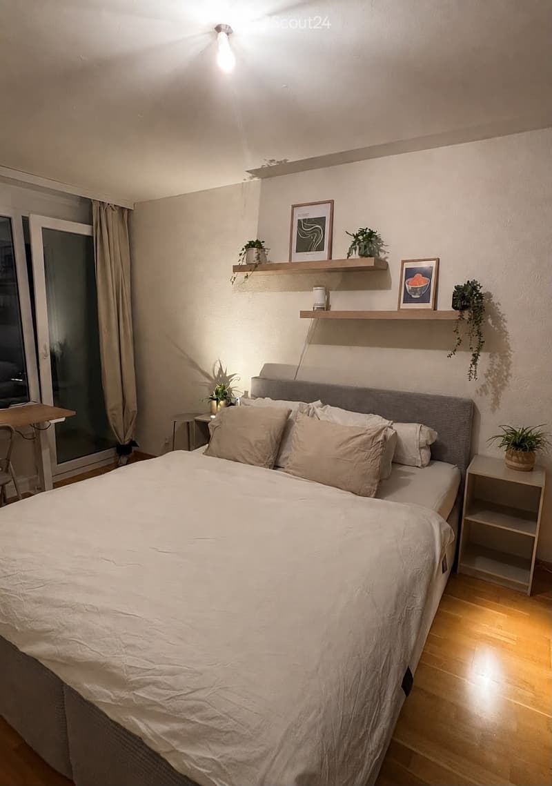 Modern bedroom with a large bed, nightstand, shelves with decor, and a small table near the window.