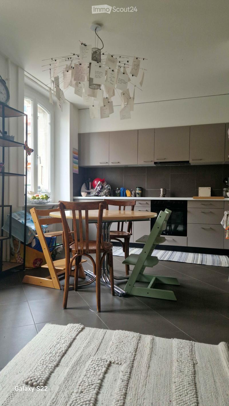 A modern kitchen with a round wooden dining table, four chairs, a green baby chair, and grey cabinets. The kitchen features a built-in oven, a refrigerator, and a countertop with various kitchen items.