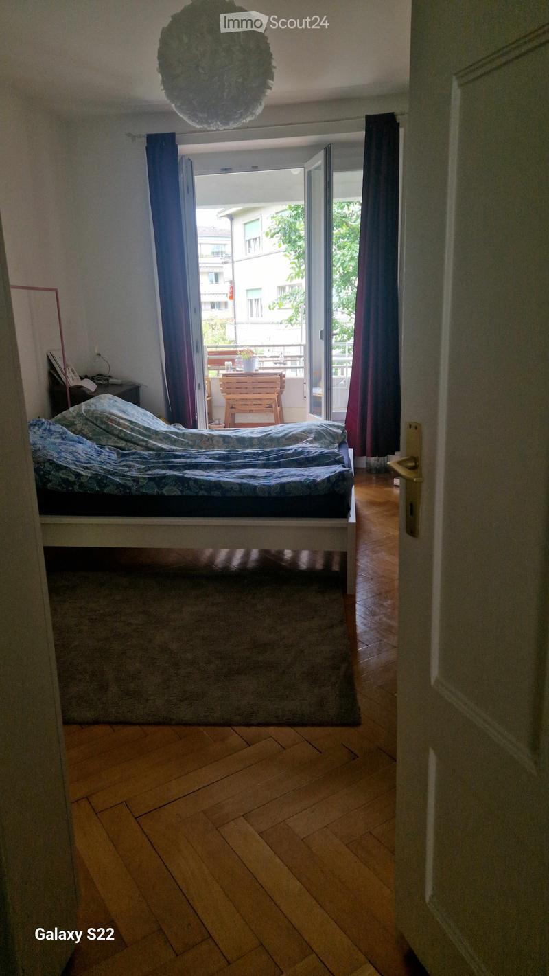 Bedroom with wooden flooring, a double bed with patterned bedding, a nightstand, and an open door leading to a balcony with outdoor furniture.