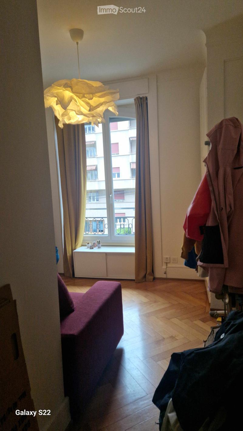 A small, well-lit living room with wooden flooring, a purple sofa, a modern lamp, a curtained window with a view of a cityscape, and a coat rack with jackets.