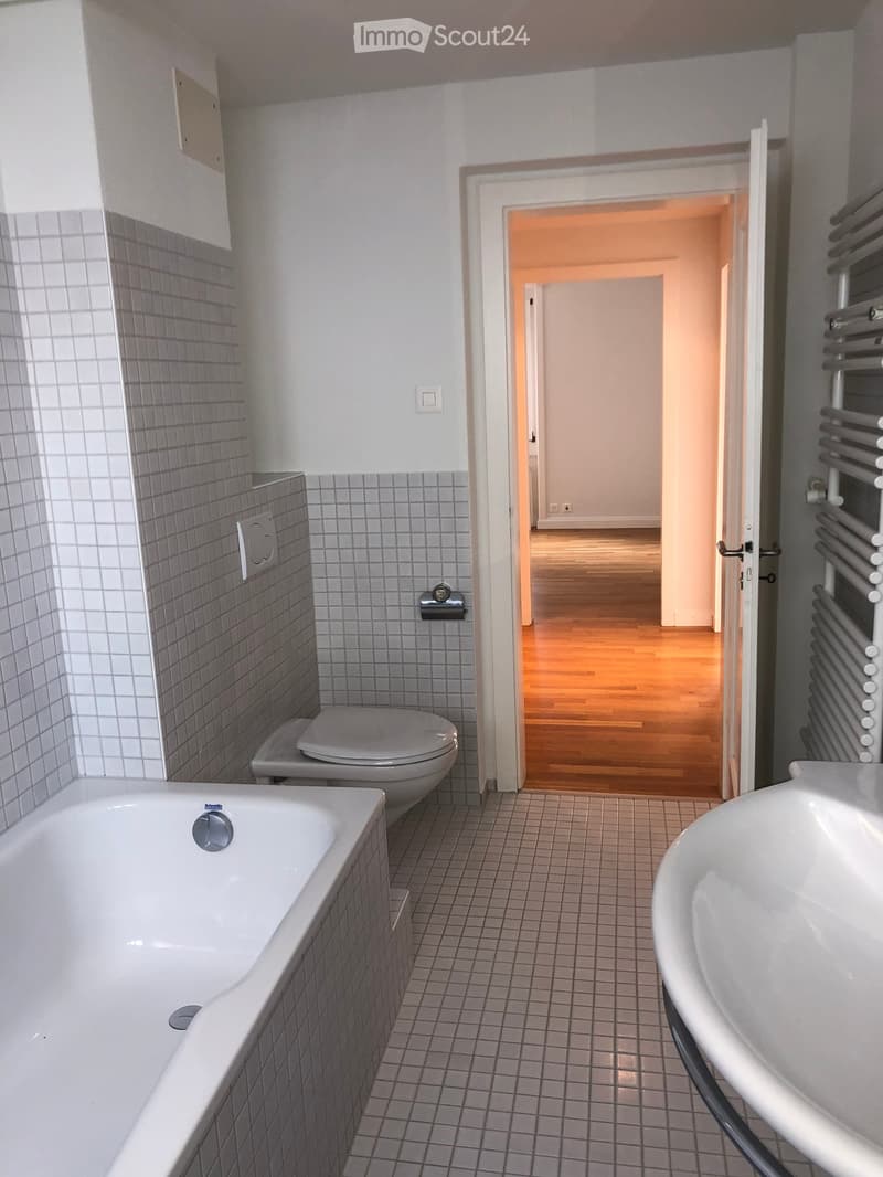 Modern bathroom with a bathtub, toilet, sink, and radiator. The walls are tiled, and there is an open doorway leading to another room with wooden flooring.