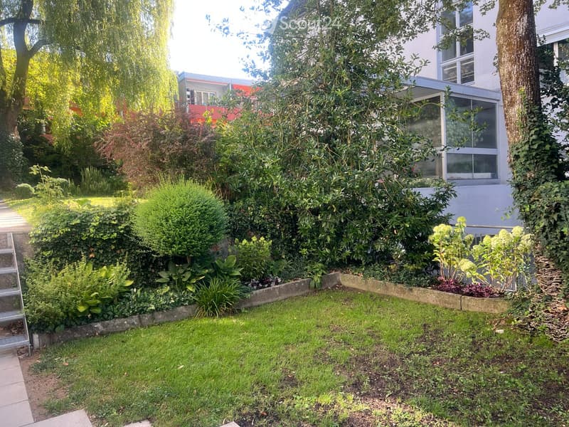 Well-maintained garden with a variety of shrubs, trees, and a neatly trimmed lawn. The garden features a raised border with plants and a modern building in the background.