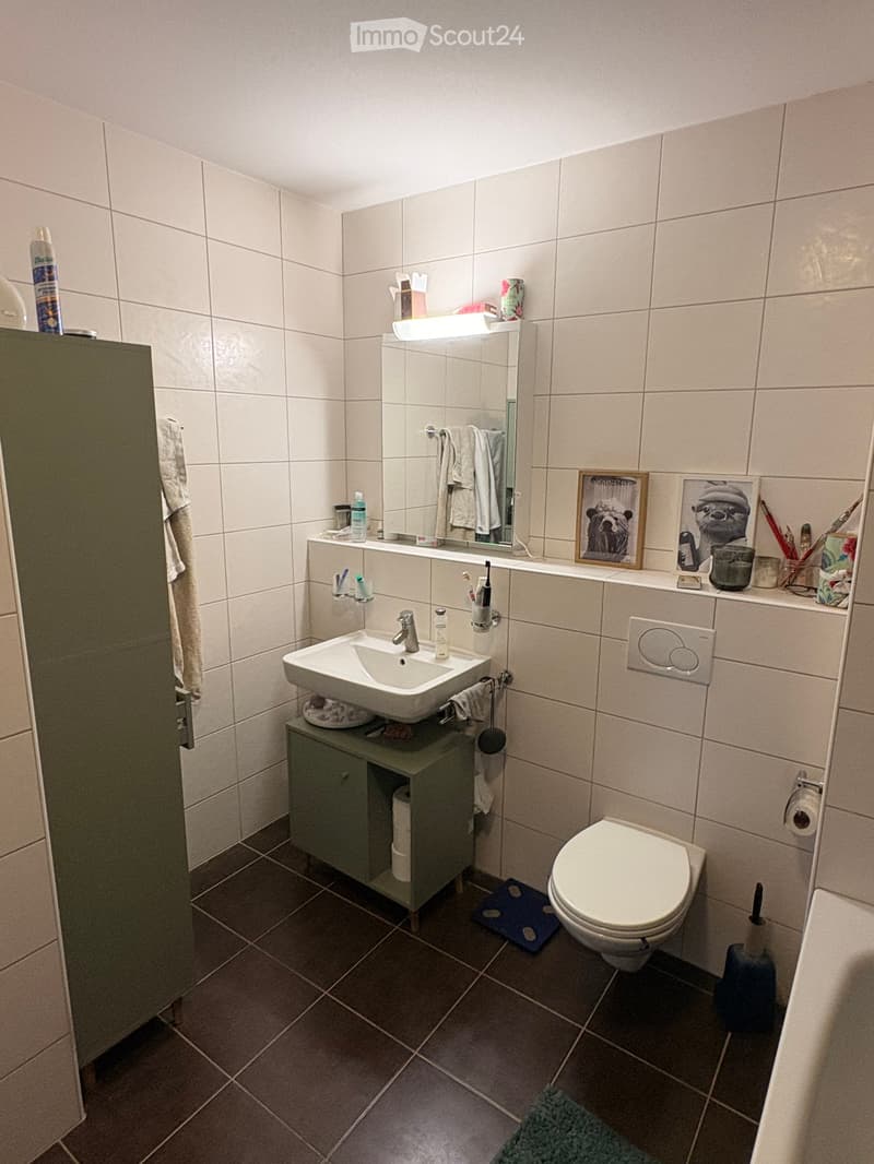 A compact bathroom with a white sink, mirror, and toilet. The sink has a modern faucet and is placed on a small green cabinet. A mirror with a built-in light is above the sink, and a towel hangs on the side. The walls are tiled, and the floor has dark til