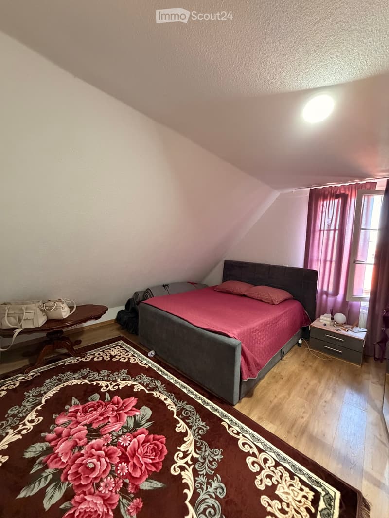Bedroom with a double bed, nightstand, small table with handbags, and a patterned rug.