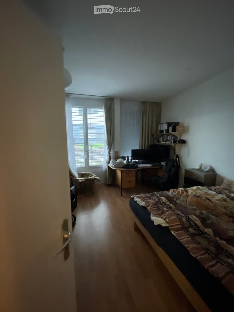 Well-lit bedroom with wooden flooring, a bed with patterned bedding, a desk with a computer, a chair, and a window with curtains.