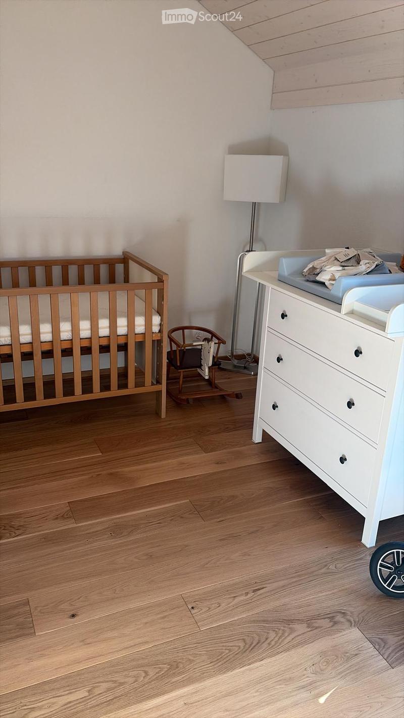 A well-lit nursery with a wooden crib, changing table, floor lamp, and a small wooden chair. The room features light wooden flooring and white walls.