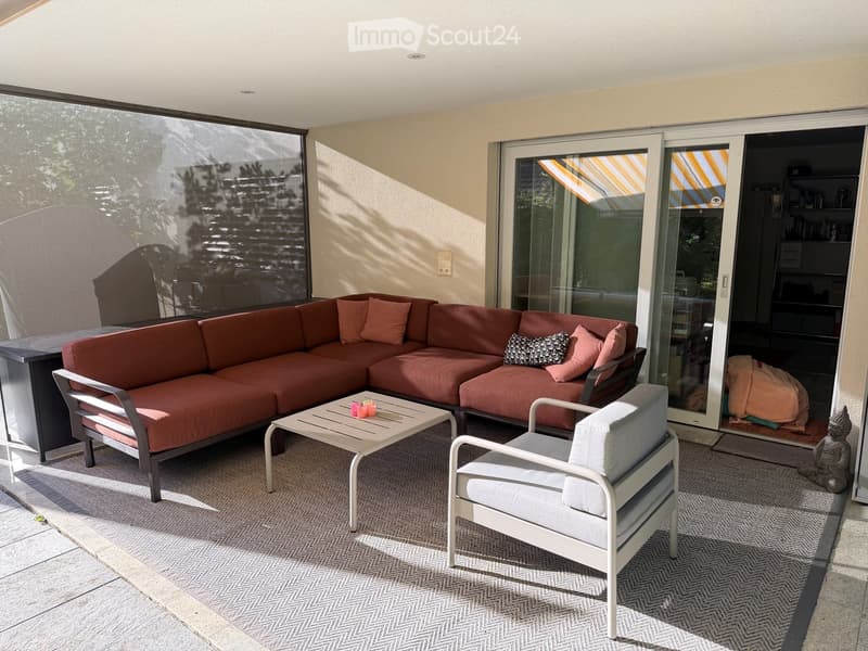 A modern outdoor patio with a red sectional sofa, white armchair, small table, and decorative pillows. Large glass doors lead to an indoor space.
