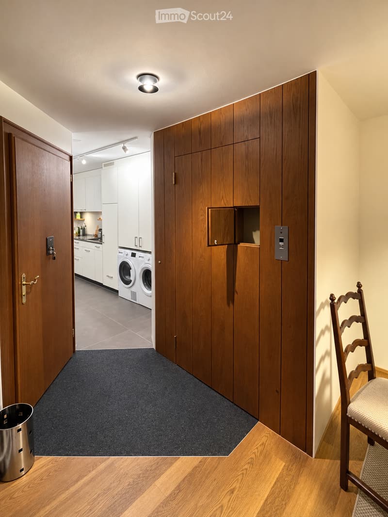 A modern hallway with wooden paneling, a closed wooden door, and a view into a kitchen area with white cabinets, a washing machine, and a dryer. A wooden chair is visible on the right side.