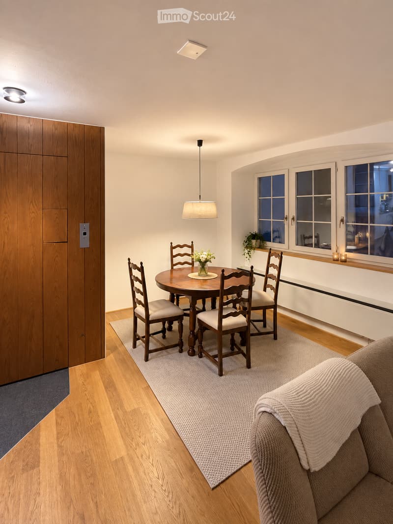 A modern dining and living area with a round wooden dining table, four chairs, a sofa, a pendant light, and a flower vase on the table. The room features wooden flooring, large windows, and a gray rug.