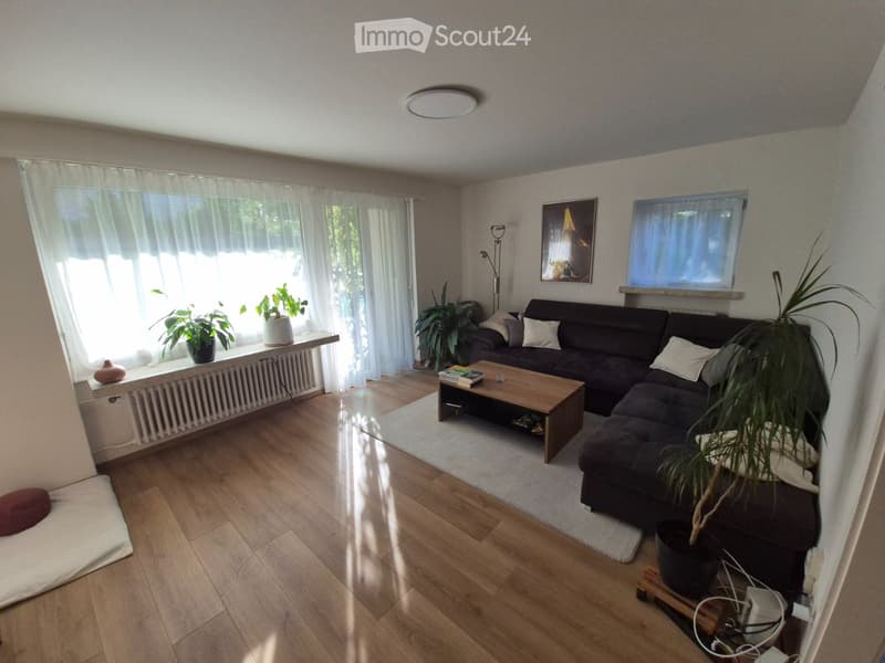 Bright living room with wooden flooring, large windows with white curtains, a dark sectional sofa, a wooden coffee table, potted plants, and a picture frame on the wall.